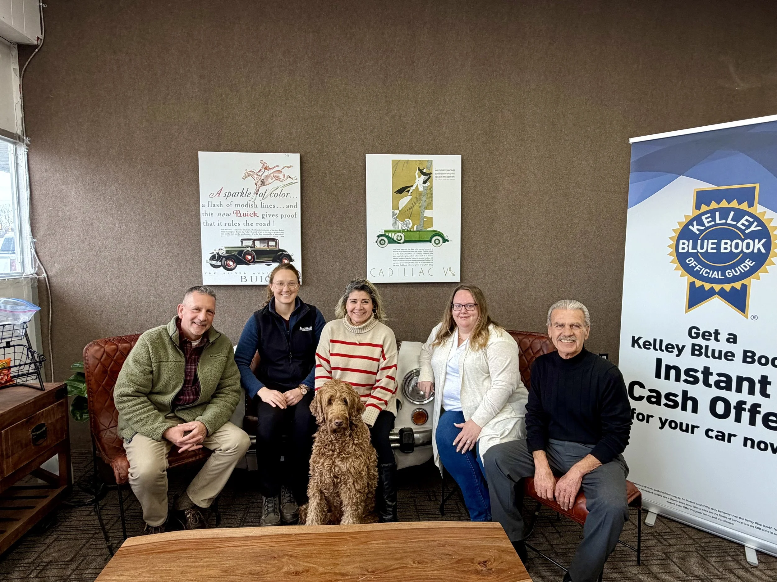 A group of six people, including one woman with glasses and a dog, sitting in an office or meeting room. Behind them are three posters, one about Buick cars, and a Kellogg's Blue Book banner on the right side.