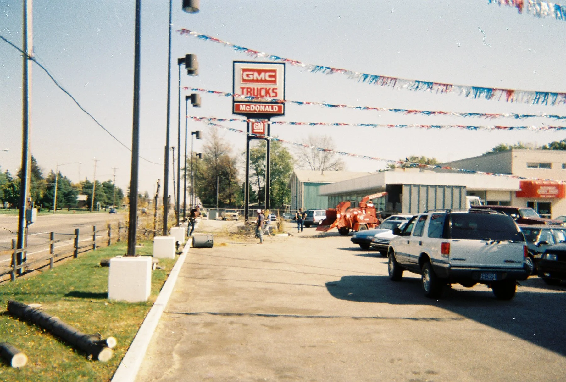 A car dealership lot decorated with red, white, and blue banner streamers. Several cars are parked, and a few people are standing around, possibly inspecting vehicles or talking. There are power poles and trees along a nearby road in the background, with a mostly clear sky overhead.