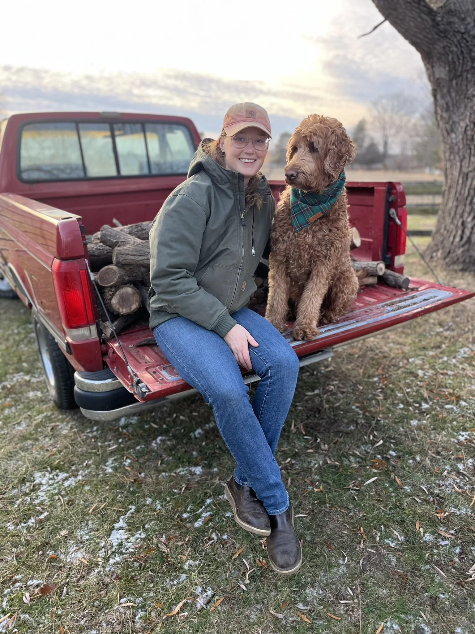 A woman smiling and wearing glasses, a pink cap, and a green jacket, sitting on the tailgate of a red pickup truck filled with logs, outdoors near a large tree, with a dog wearing a plaid bandana sitting beside her.