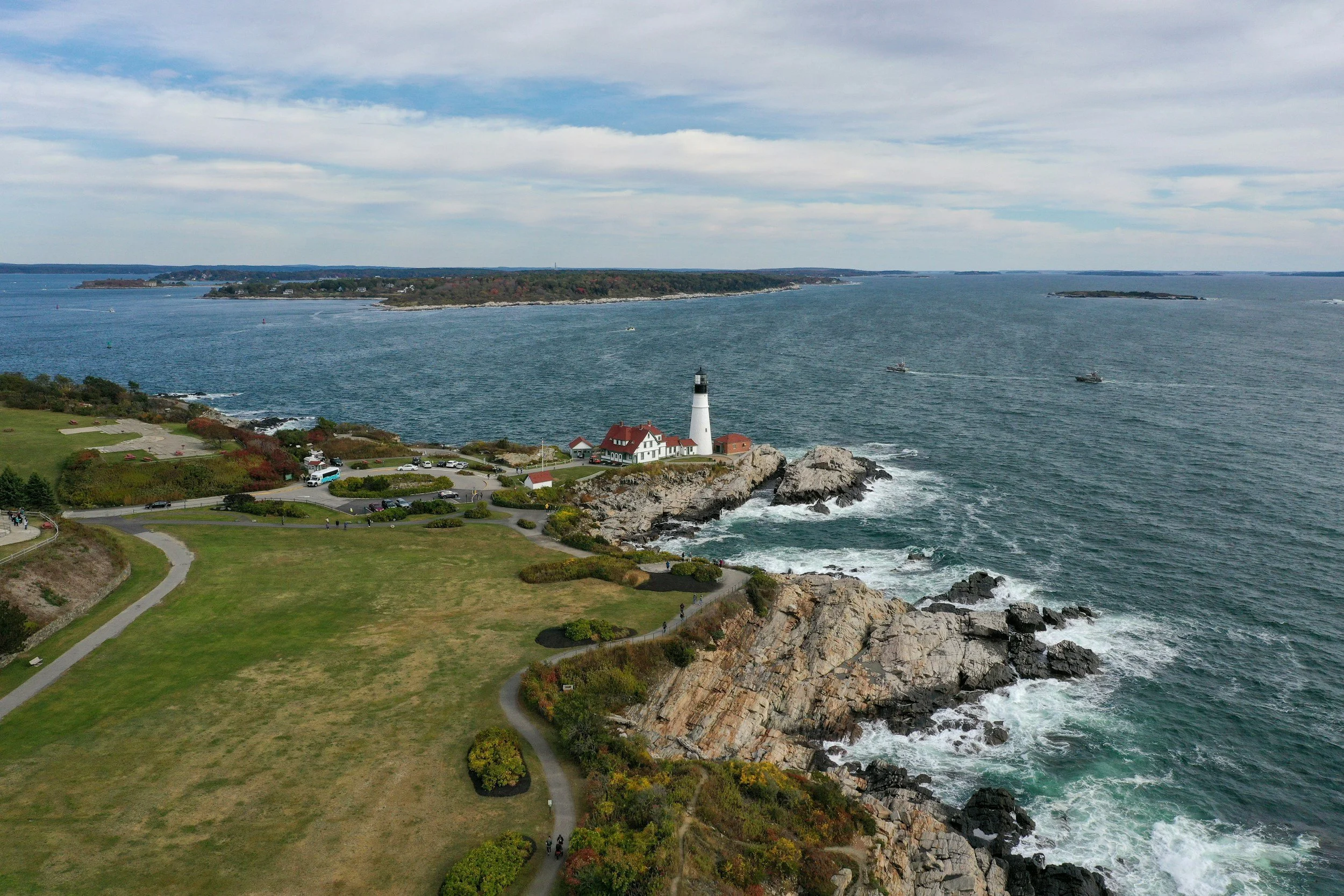 A scenic coastal view showing a lighthouse on a rocky promontory, surrounded by a park with walking paths, and boats navigating on the water. There are distant islands and a cloudy sky.