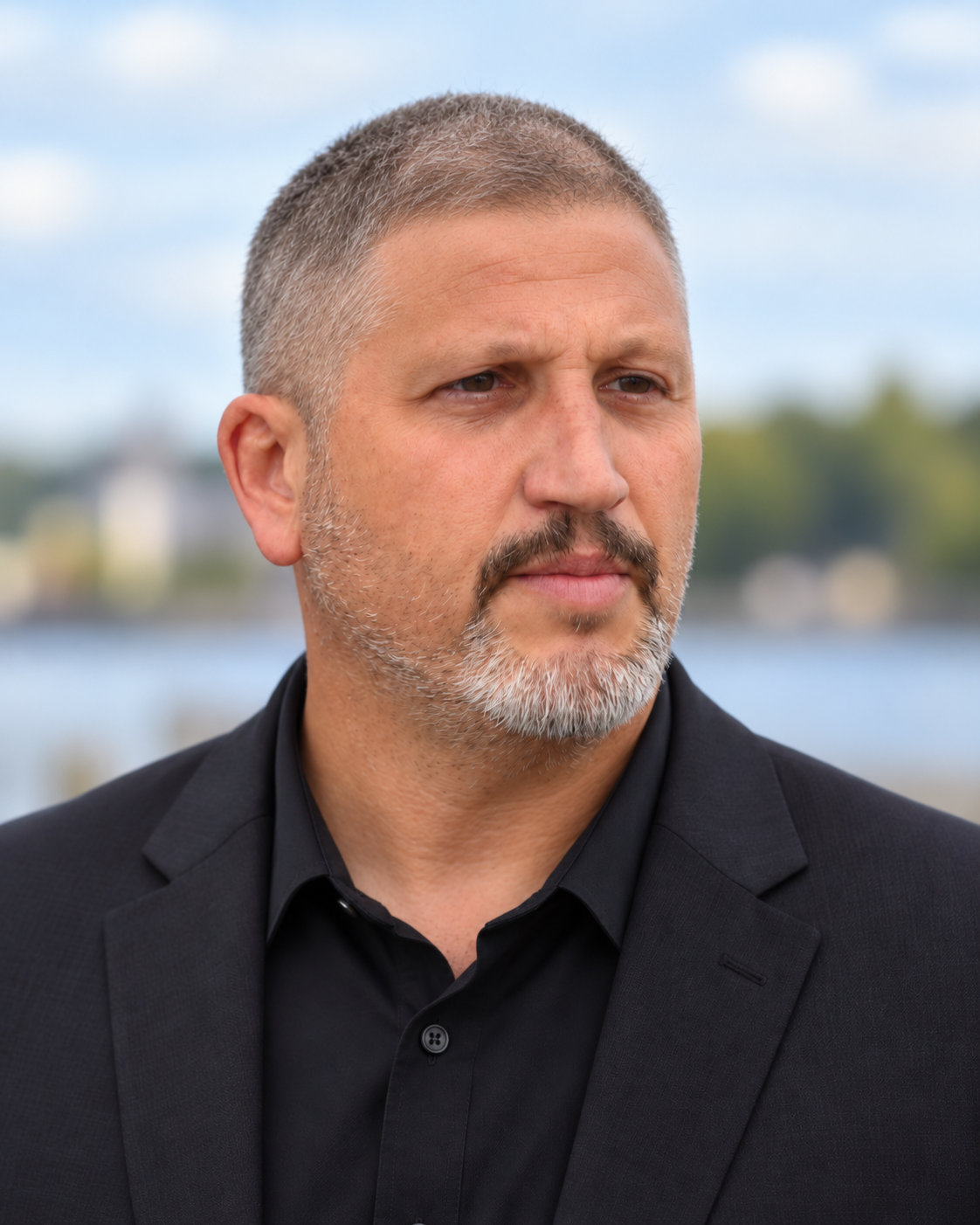 A middle-aged man with short gray hair and a beard wearing a black suit and shirt outdoors near water with a blurred landscape in the background.