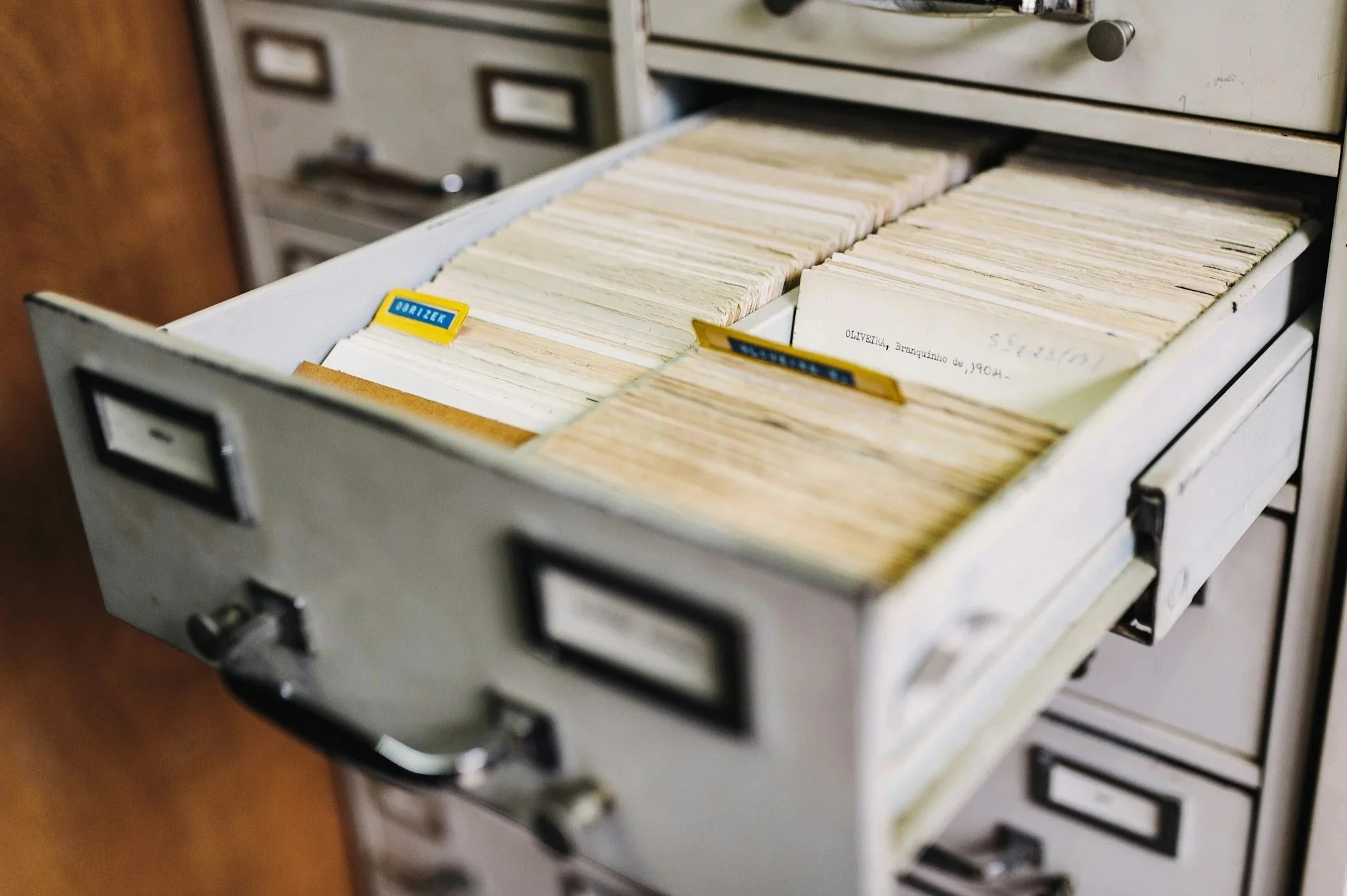 A close-up view of a vintage metal filing cabinet drawer filled with old paper documents organized in rows with yellow and blue labeling tabs.