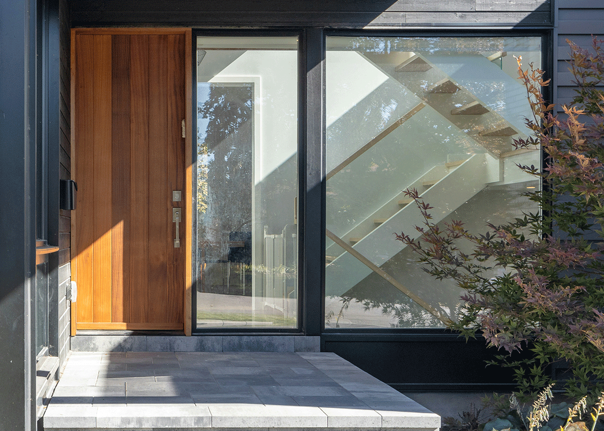 Close-up of modern house entrance with a wooden door, glass side panel, and a staircase visible through the glass, surrounded by plants.