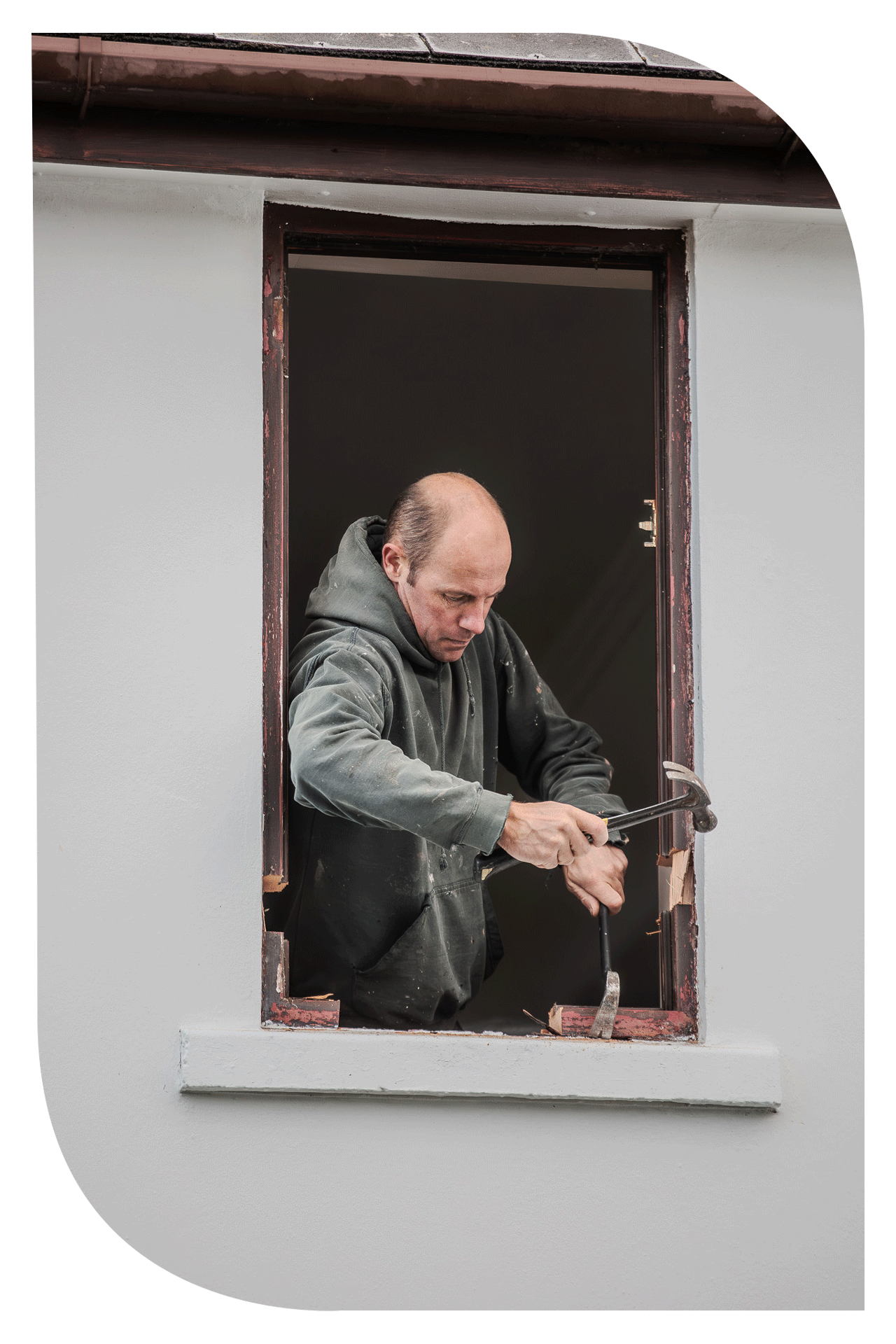 A man working on a window frame seen from outside, using a hammer and chisel, surrounded by white wall with some parts of the window frame worn and under repair.