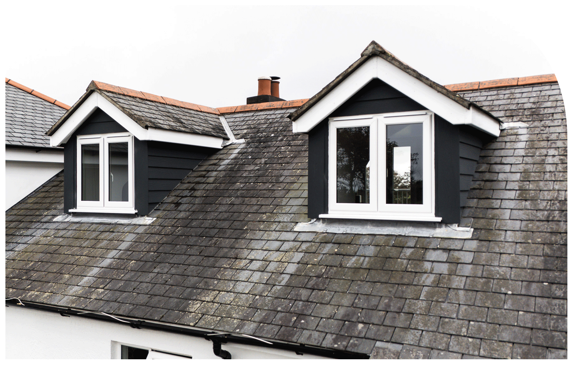 Close-up of a dark gray sloped rooftop with two dormer windows and a chimney, weathered shingles, and moss patches.