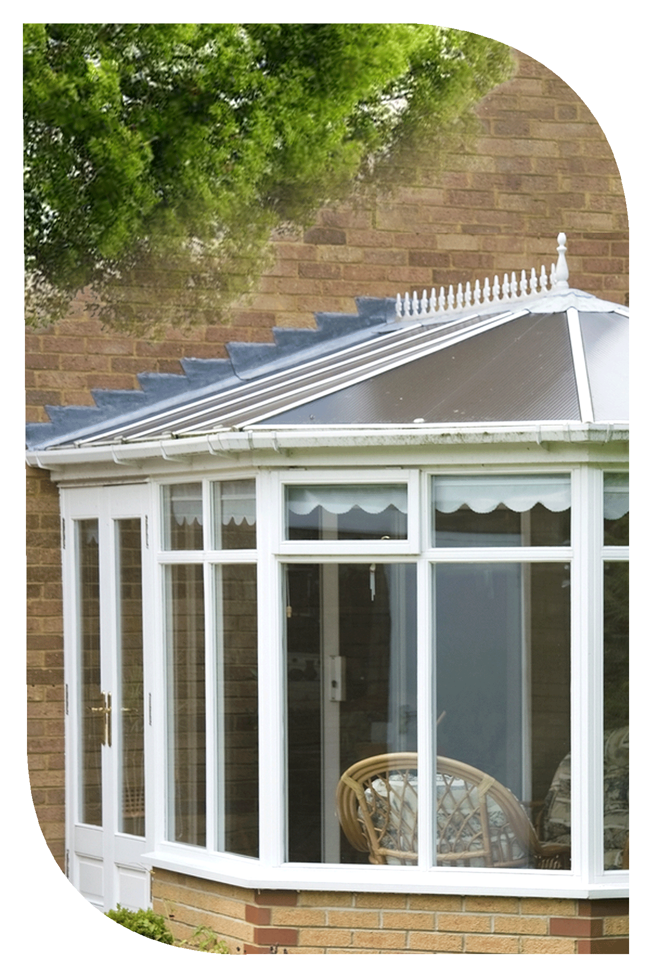 A sunroom with white framed bay windows and a glass roof, attached to a brick house. A wicker chair is visible inside the sunroom.