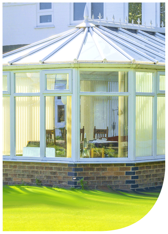 Sunroom with glass walls and a metal roof, attached to a white house, surrounded by a green lawn.