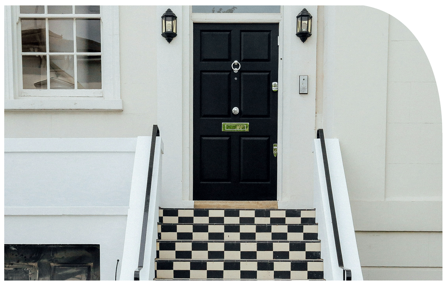 Front door with black panels, flanked by two wall-mounted lantern lights, accessed by a black and white checkered tile staircase with white railings, in a white building with a window on the left.