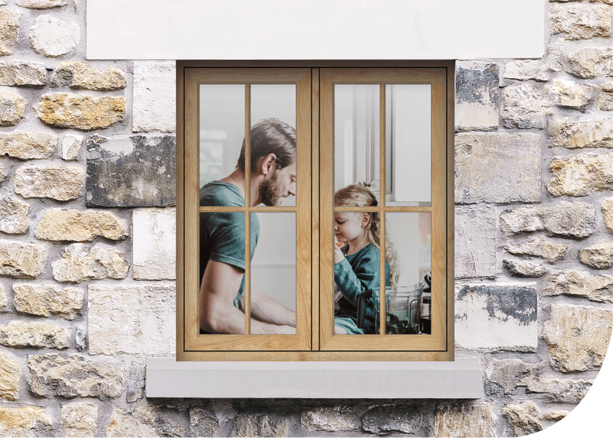 A man and a young girl are sitting at a table inside, seen through a window with wooden frames set in a stone wall exterior. The man has a beard and is wearing a teal shirt, and the girl has curly hair and is wearing a teal long-sleeve shirt. They are facing each other, with the girl touching her chin and the man looking down, engaged in conversation.