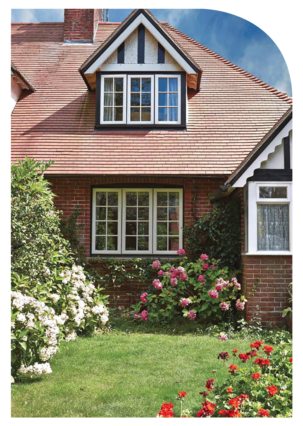 A traditional brick house with a sloped shingled roof, featuring a central upper dormer window with white trim, surrounded by flower bushes and a green lawn under a clear blue sky.
