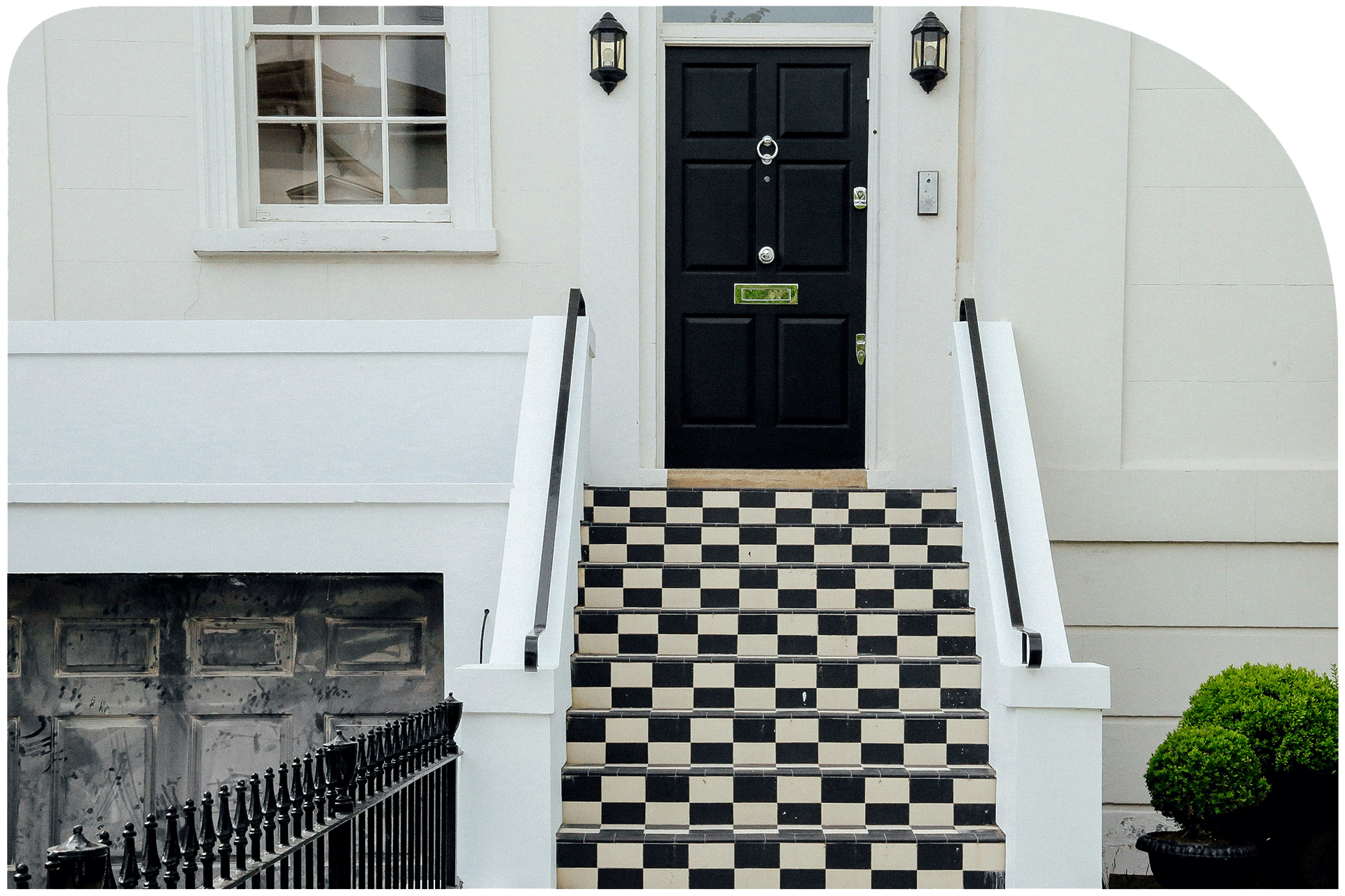 Front porch of a house with black front door, white stairs with black and white checkered tiles, two black lantern-style outdoor lights, a small side window, and neatly trimmed bushes.