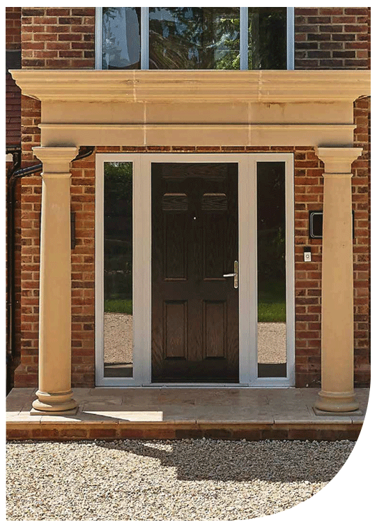Front view of a house entrance with a black door, flanked by sidelights, and supported by two beige stone columns under a decorative stone lintel, brick exterior walls, and a glass transom window above.