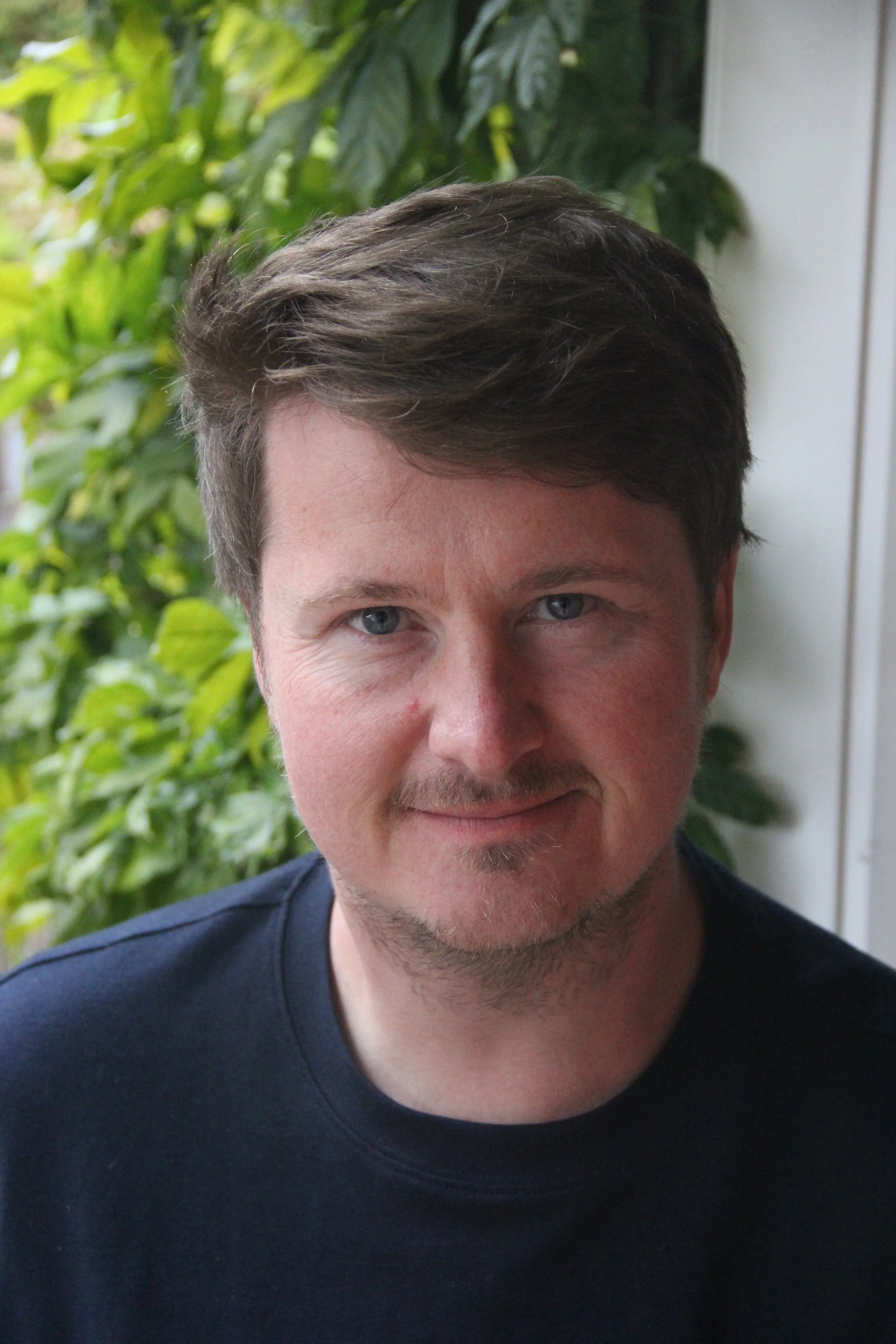 Close-up of a man with light skin, short brown hair, blue eyes, and a slight smile, wearing a black shirt, with green leafy plants and a white wall in the background.