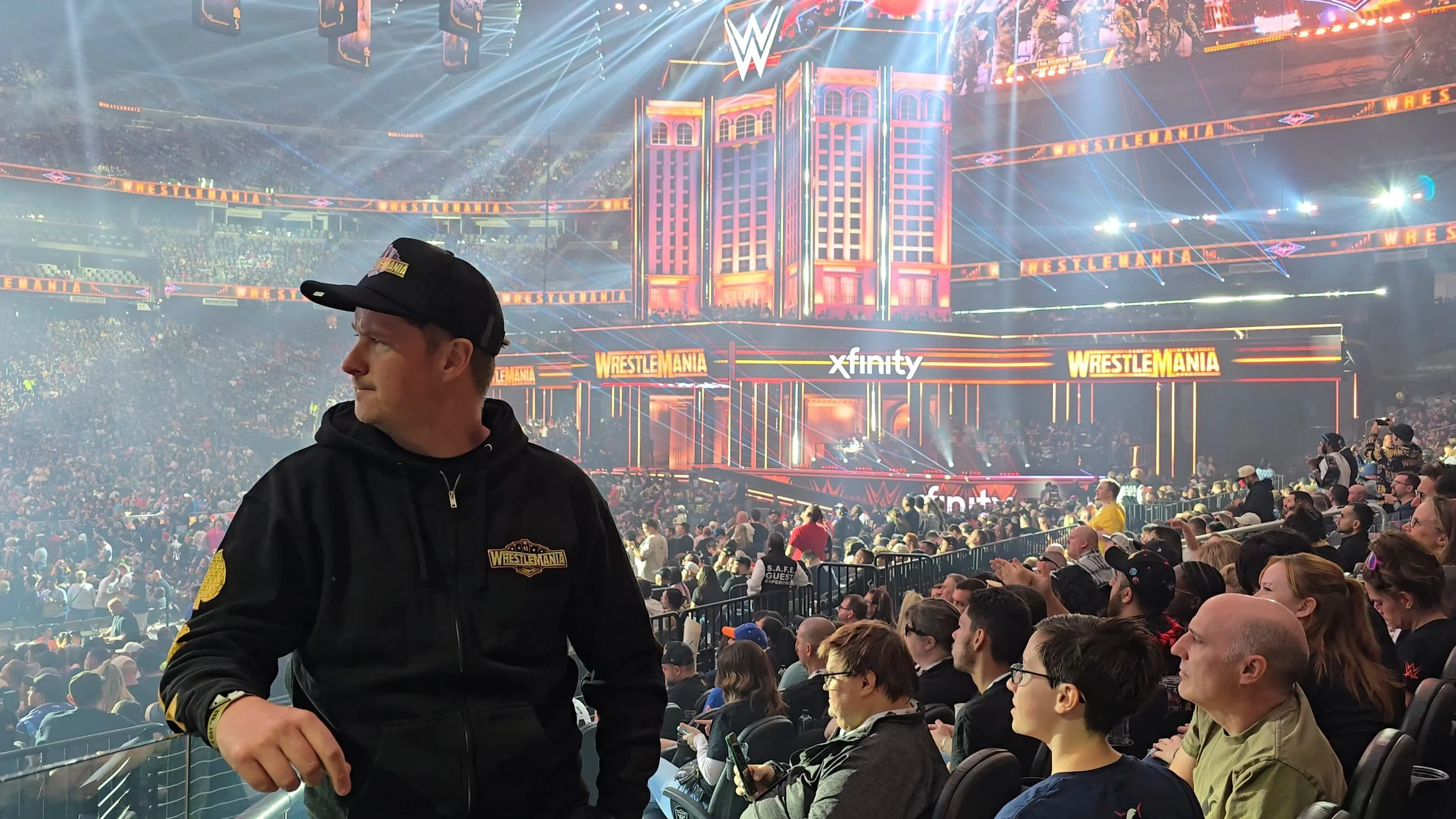 Man wearing a black WrestleMania hoodie and cap, standing in a crowded arena during a WWE event with bright stage lights, large screens displaying WrestleMania branding, and a large audience.