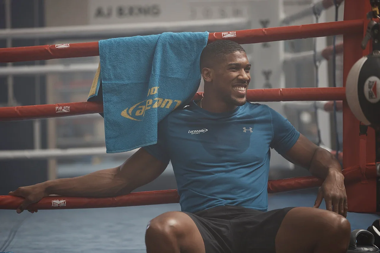 A man sitting in a boxing gym, leaning against red boxing ring ropes, smiling, with a blue towel hanging over the ropes behind him, wearing a blue athletic shirt and black shorts.