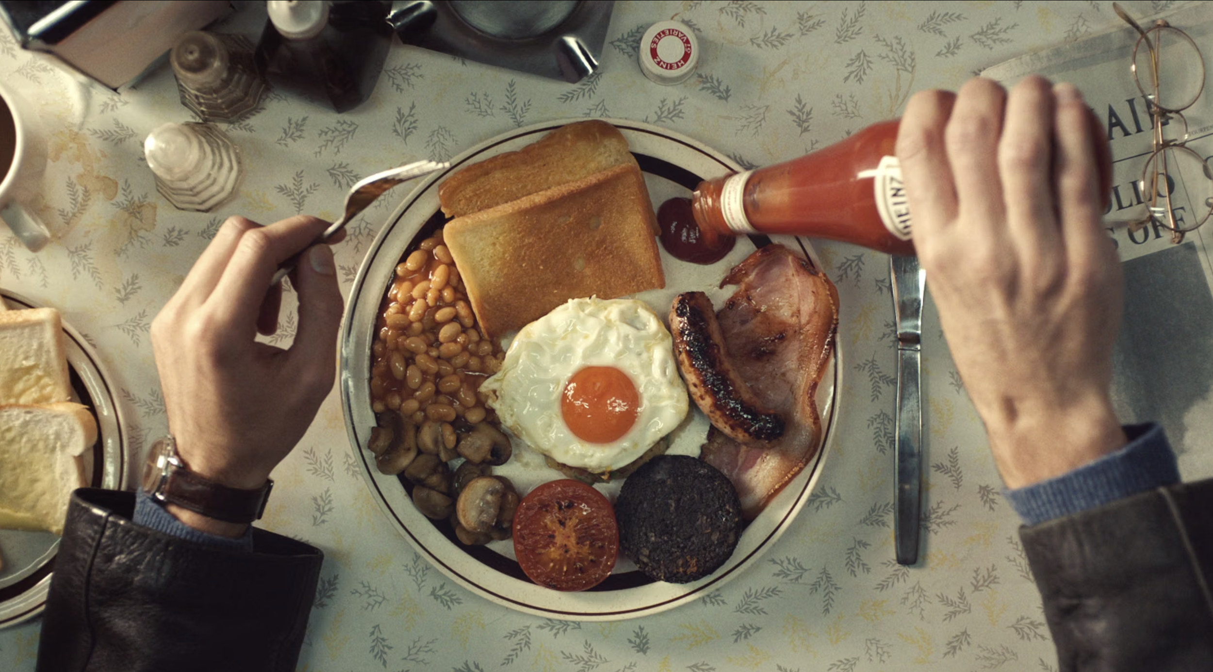 Overhead view of a breakfast plate with fried egg, bacon, baked beans, grilled tomato, mushroom, and black pudding, surrounded by breakfast items and condiments on a festive tablecloth.