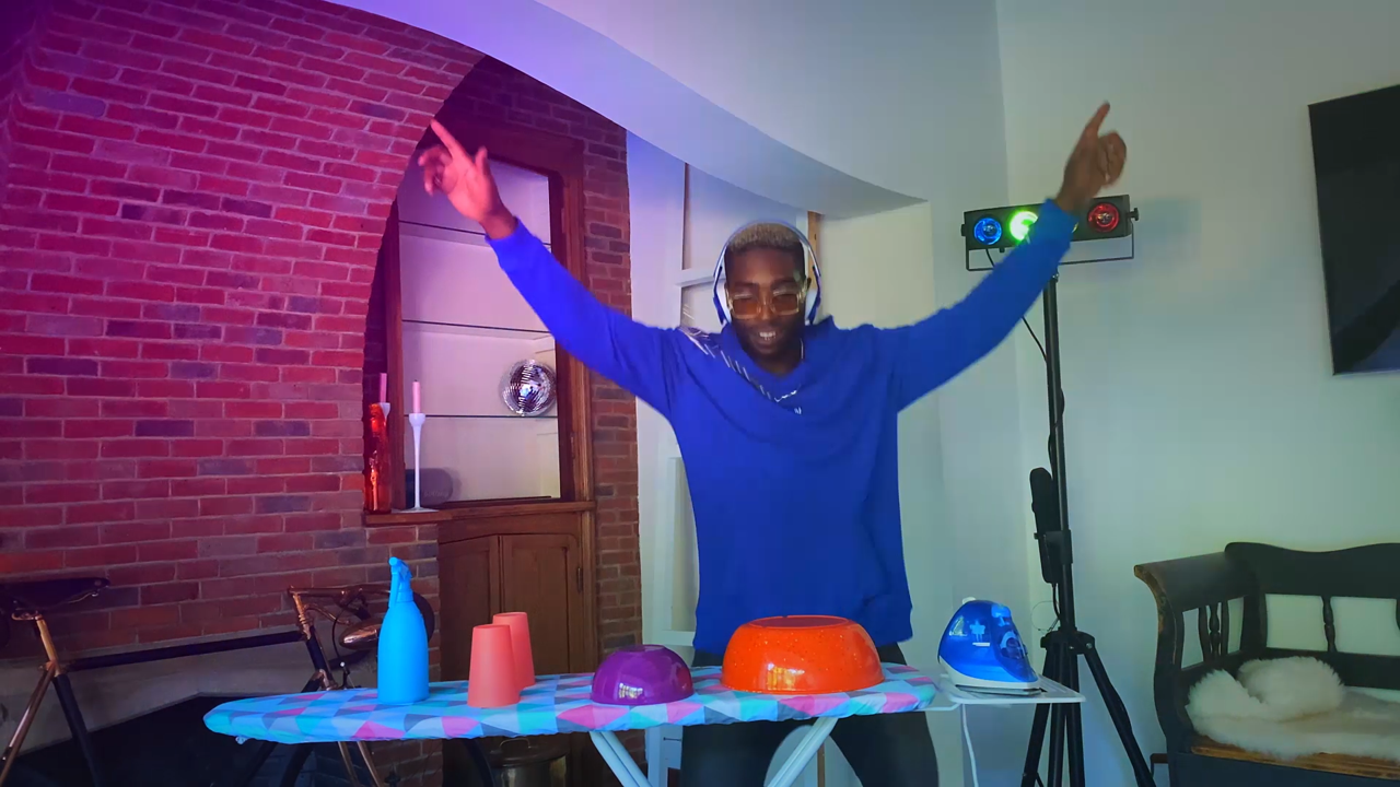 Person DJing at a party, wearing headphones, with arms raised, using a colorful table with bowls and cups, in a room with a brick wall and disco lights.