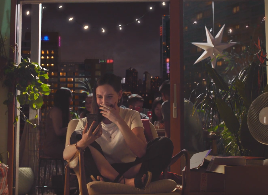 A woman sitting on a chair indoors, looking at her phone with a surprised or amused expression, with a city skyline visible through the window behind her, decorated with string lights and a large star-shaped ornament.