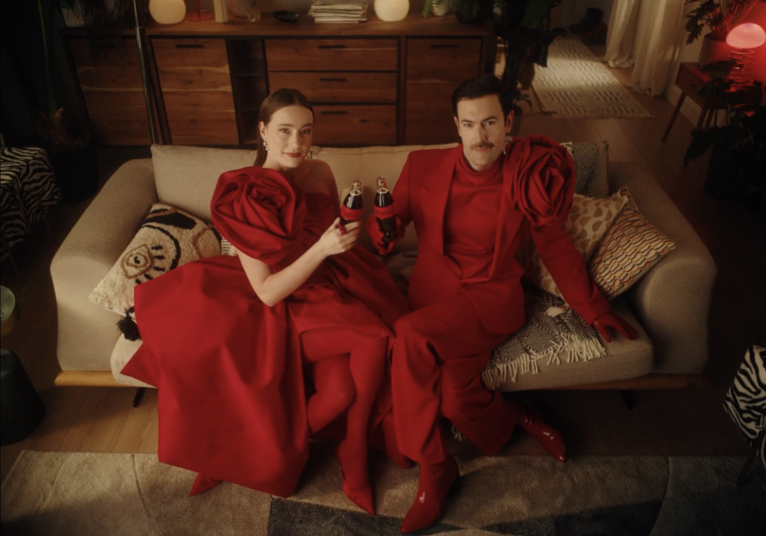 A man and woman sitting on a sofa, dressed in red outfits with large, puffy sleeves, holding small bottles of Coca-Cola, in a warmly lit living room decorated with plants and cushions.
