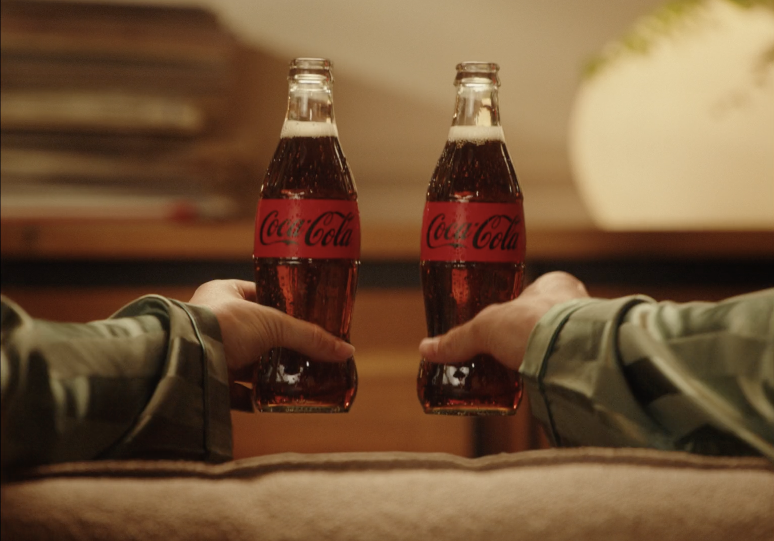 Two hands holding Coca-Cola glass bottles clinking together, seen from below.