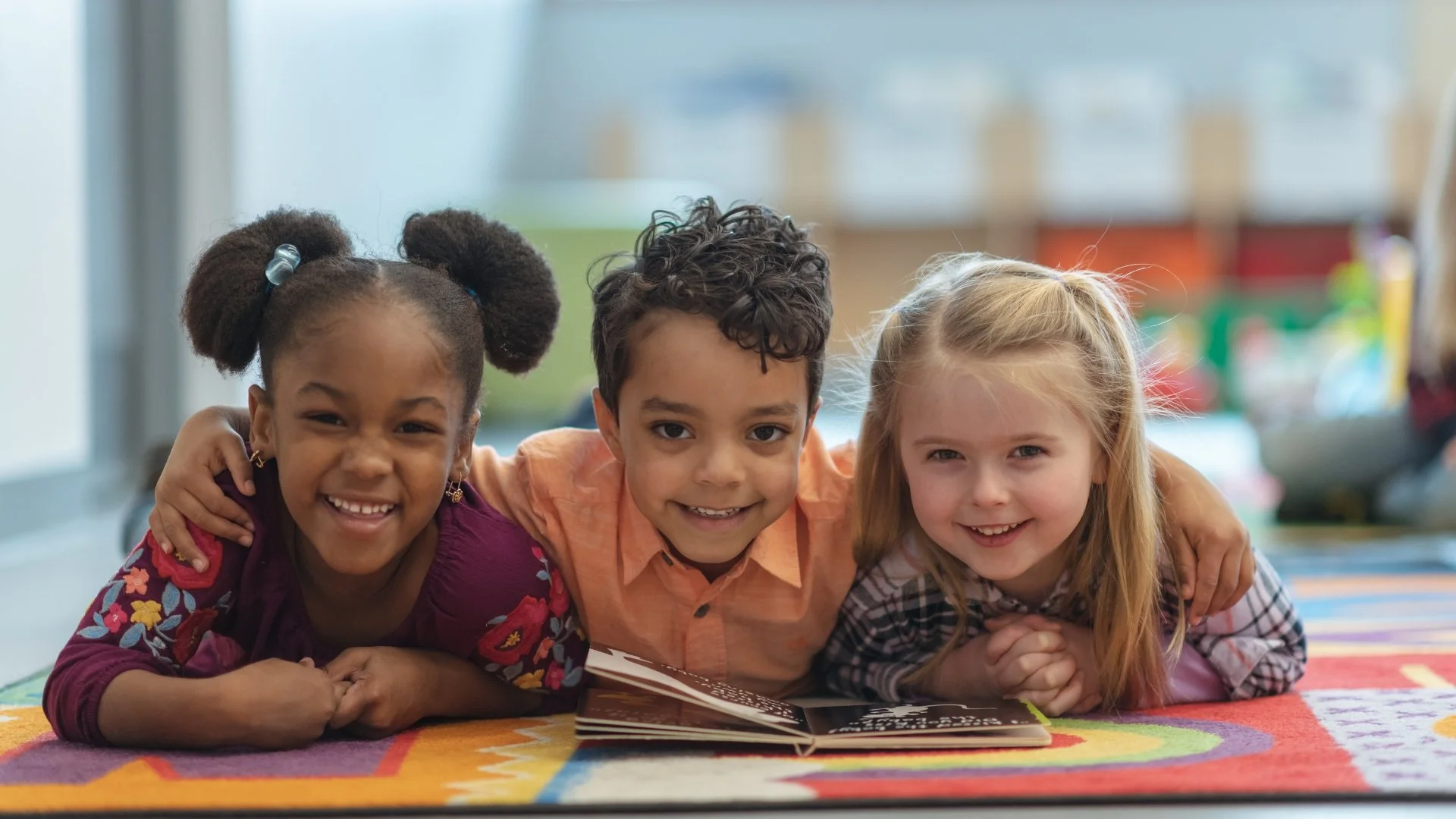 Three smiling children reading an open book together on a colorful classroom rug