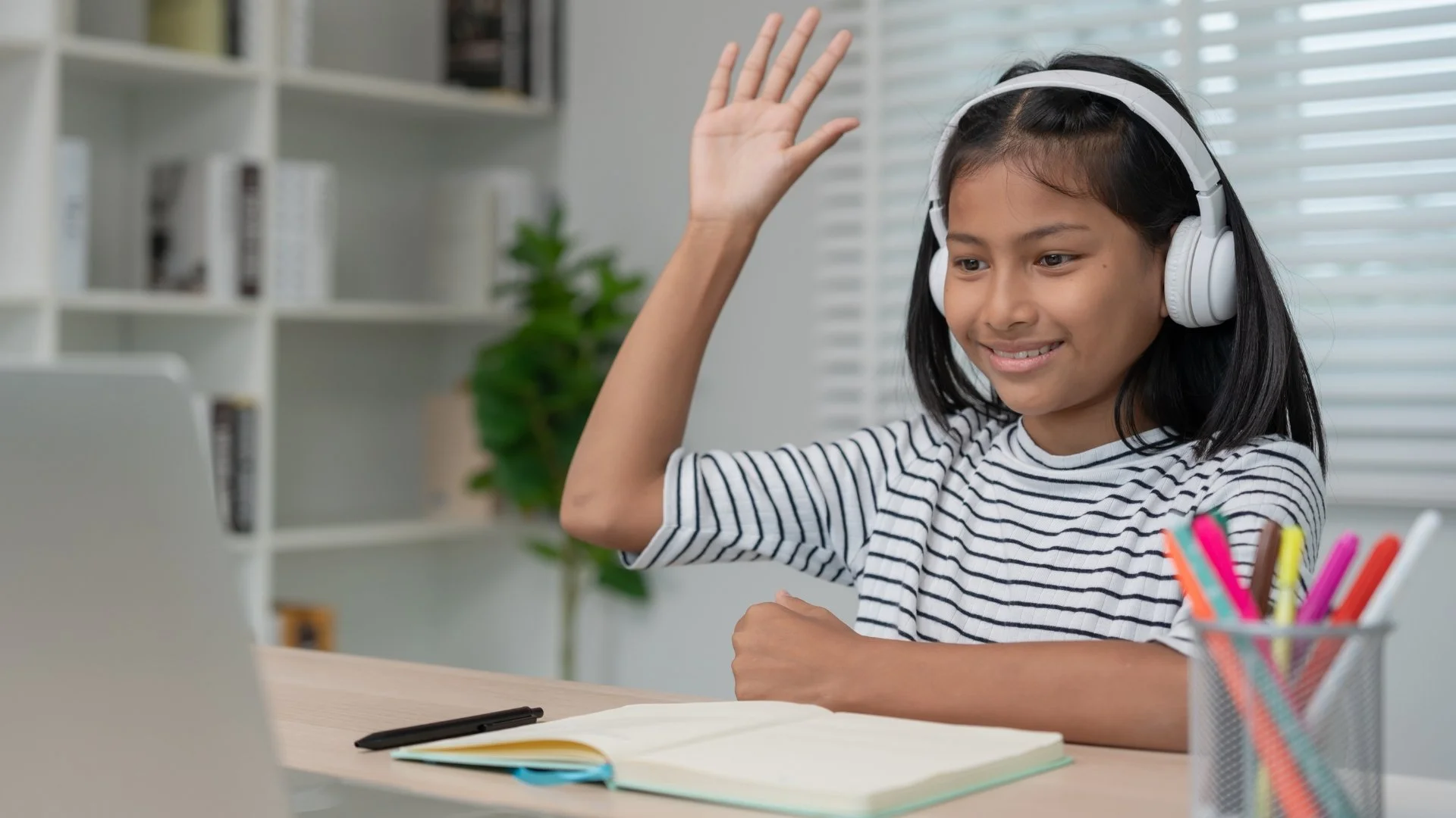 Smiling student wearing headphones raises her hand during an online class at a desk with an open notebook and laptop.
