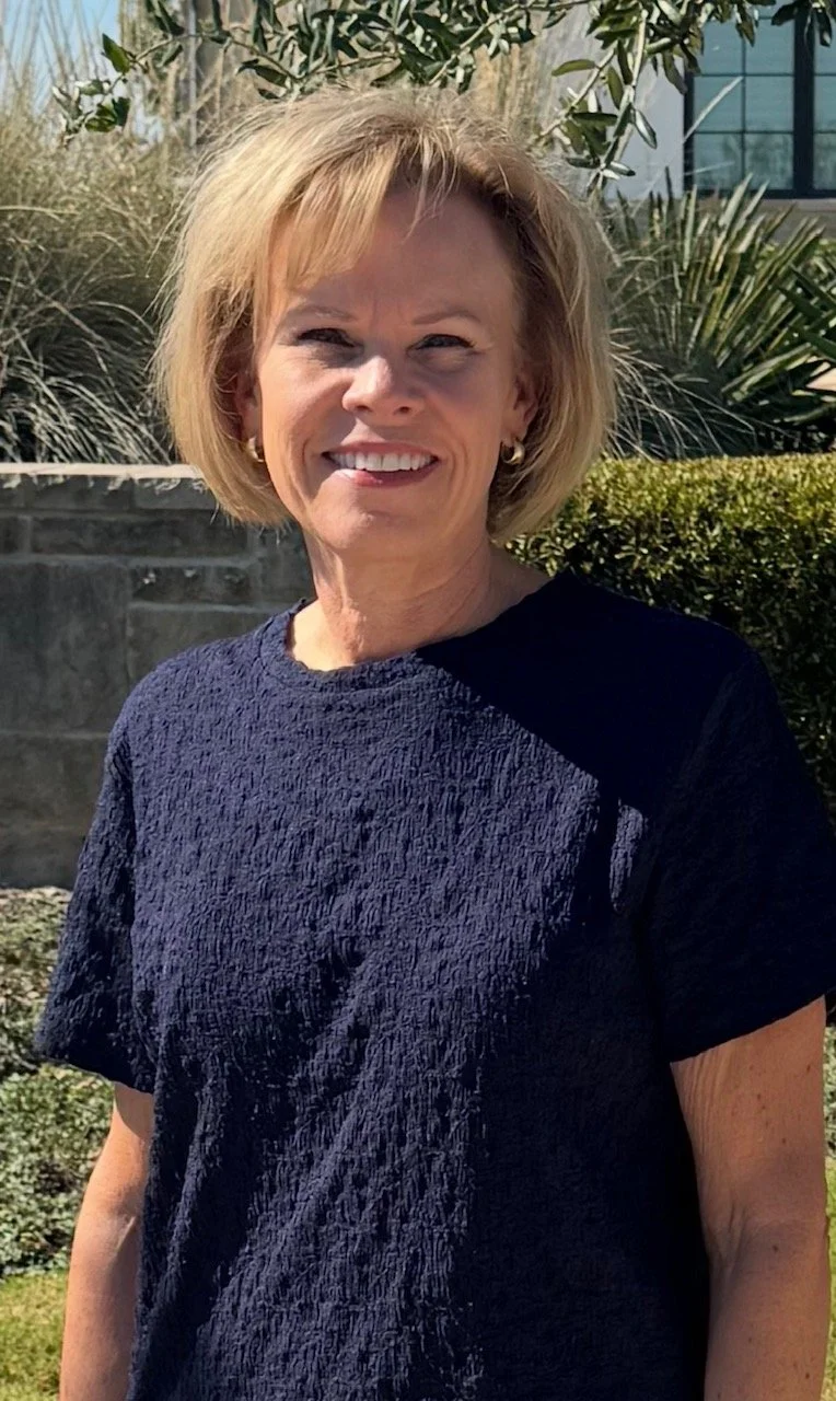 Smiling blonde woman in a navy blue top standing outdoors in a landscaped garden.