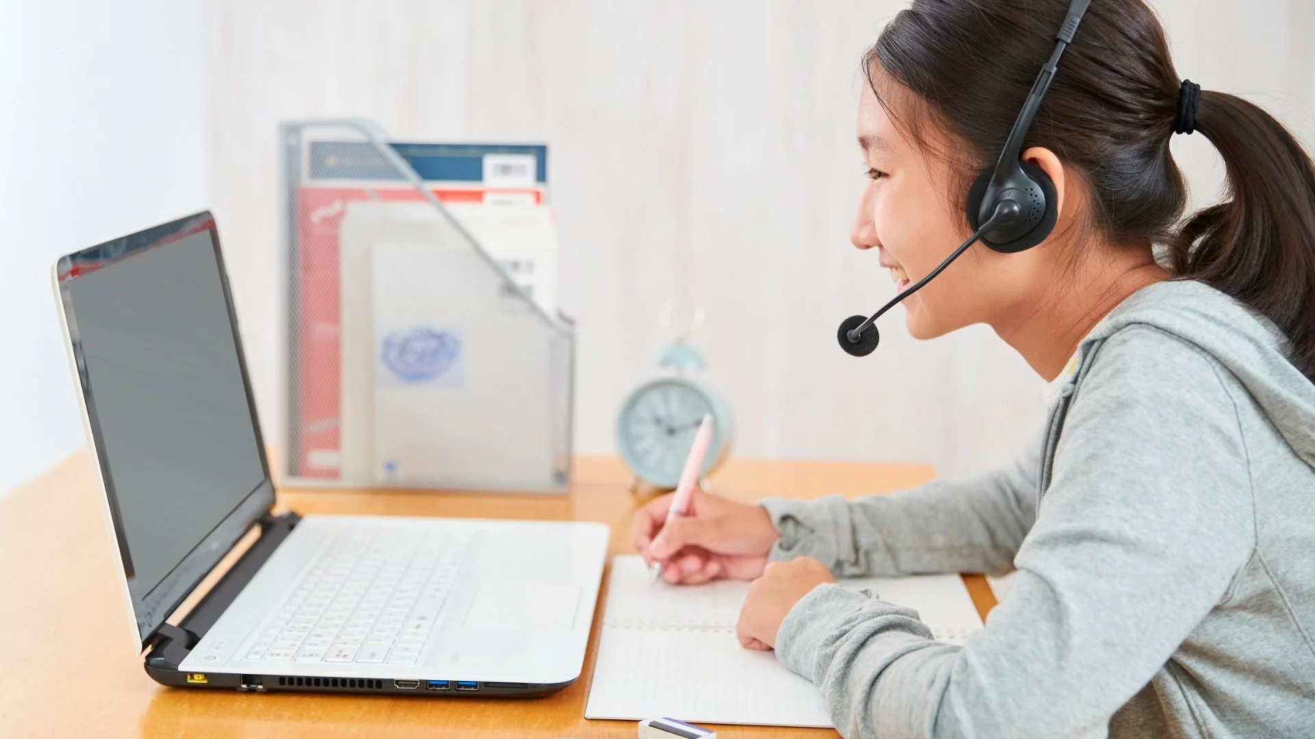 Woman wearing a headset writes notes beside an open laptop at a desk.