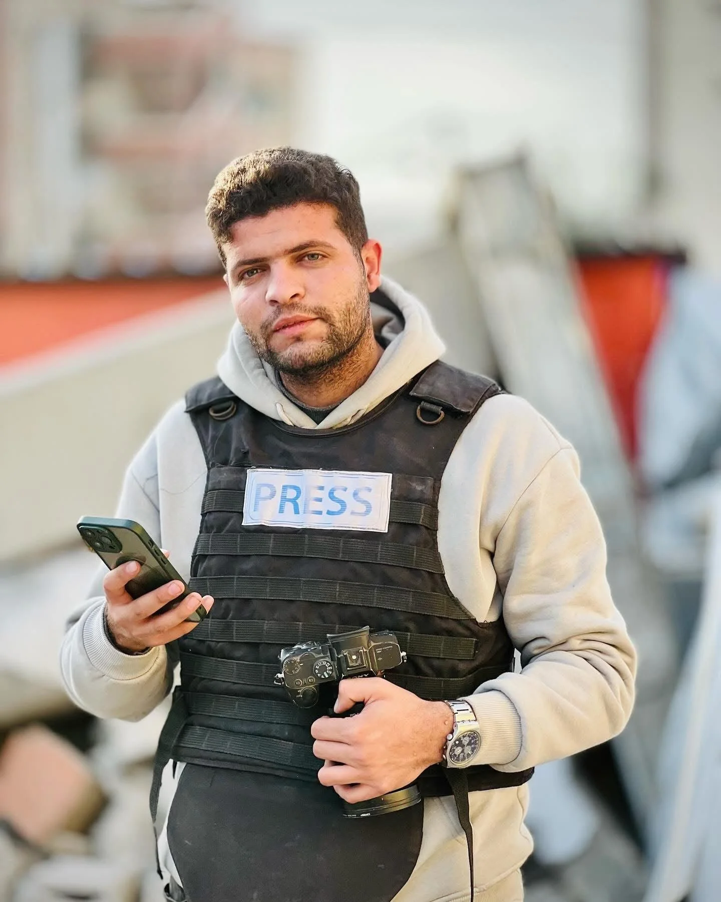 A man wearing a press badge, black vest, beige hoodie, and watch, holding a smartphone and a camera, standing outdoors with blurred background.