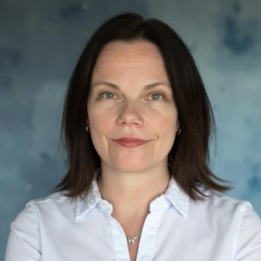 A woman with brown hair and light skin, wearing a white button-up shirt, looking at the camera with a slight smile, against a blurry blue background.