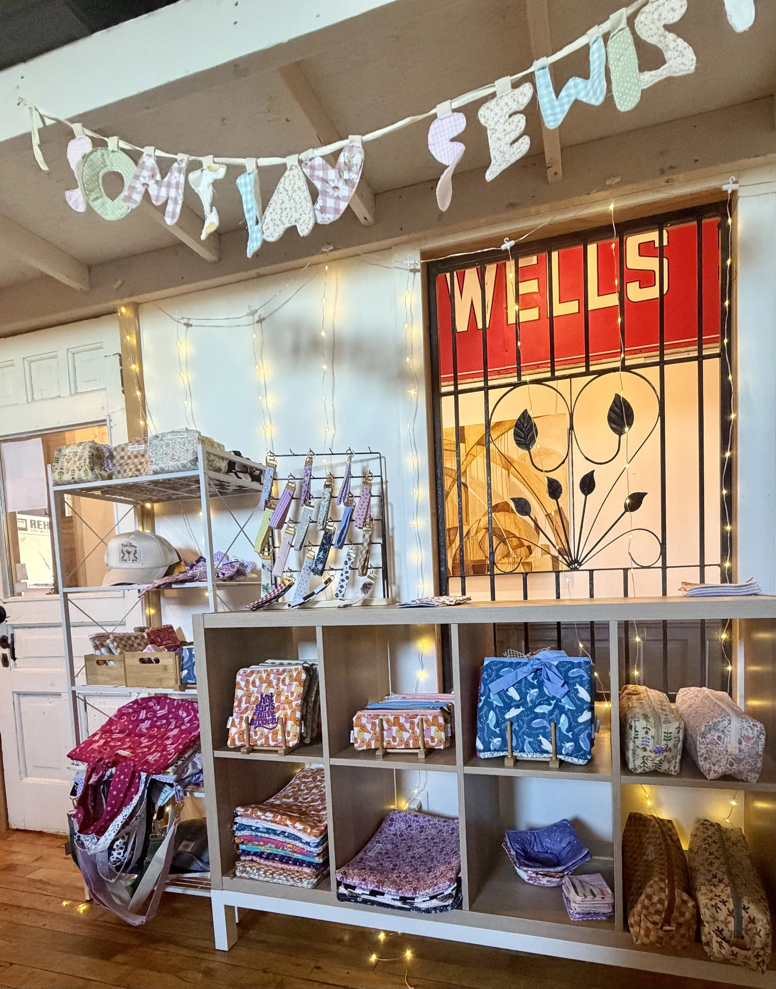 A display of fabric and clothing items on white shelves, with a decorative 'Welcome' sign hanging above and fairy lights arranged on the wall, in a cozy shop setting.
