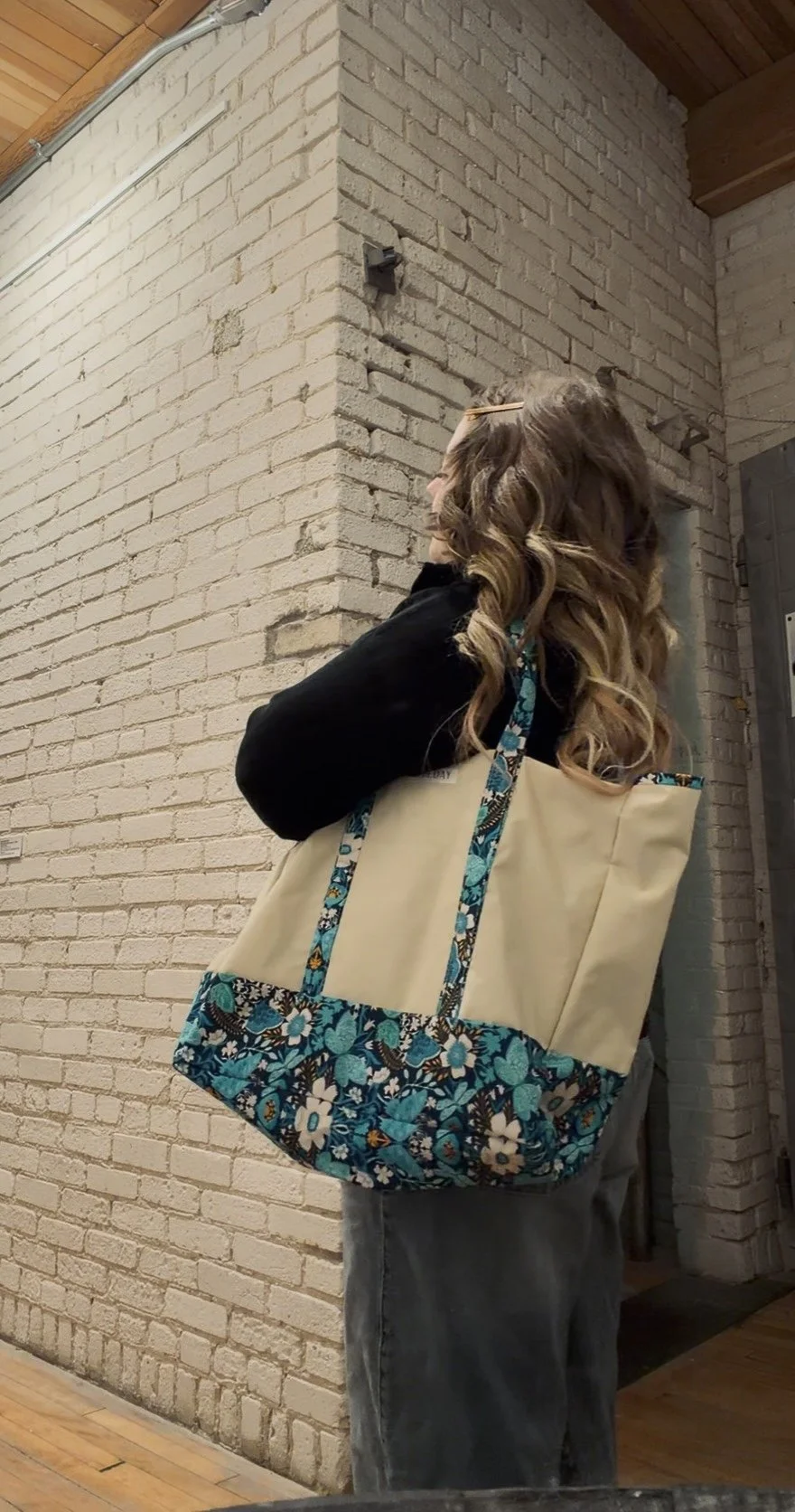 A woman with curly brown hair, wearing a black top and gray jeans, standing against a white brick wall. She is holding her head with her right hand and has a large beige tote bag with blue floral accents hanging from her shoulder.