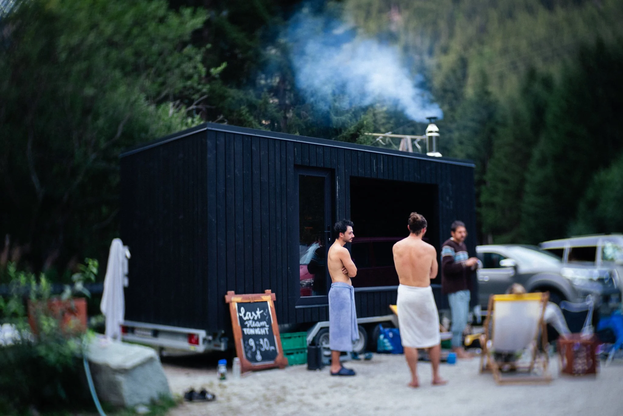 People standing near a black mobile sauna with a signboard outside that reads 'last steam today 9:30'. The background includes green trees and parked cars.