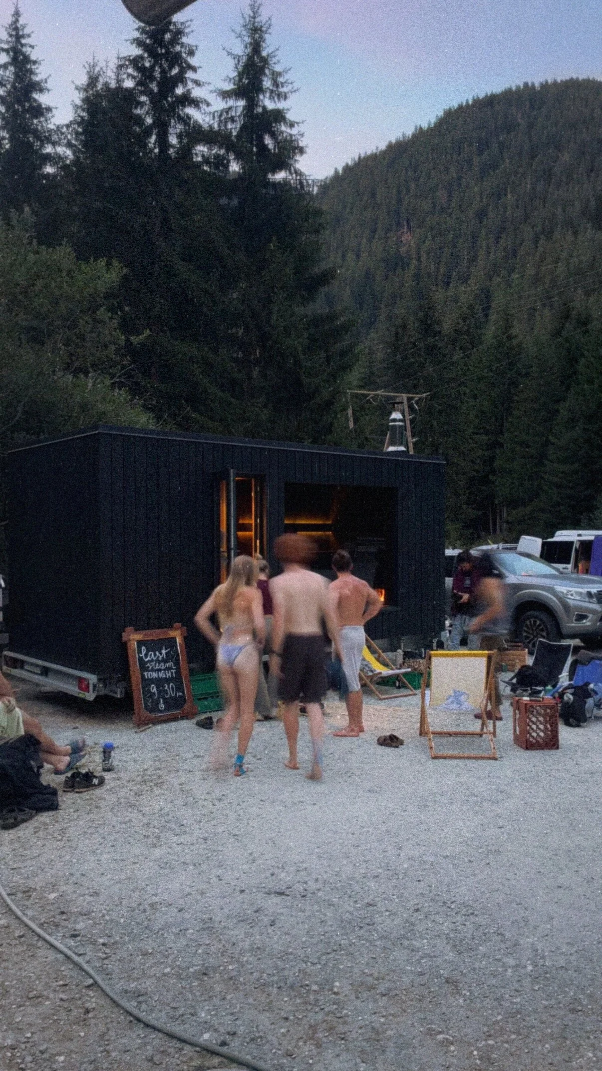 People in swimsuits gathering outside a mobile sauna with a chalkboard sign, mountain trees in the background, and cars parked nearby.
