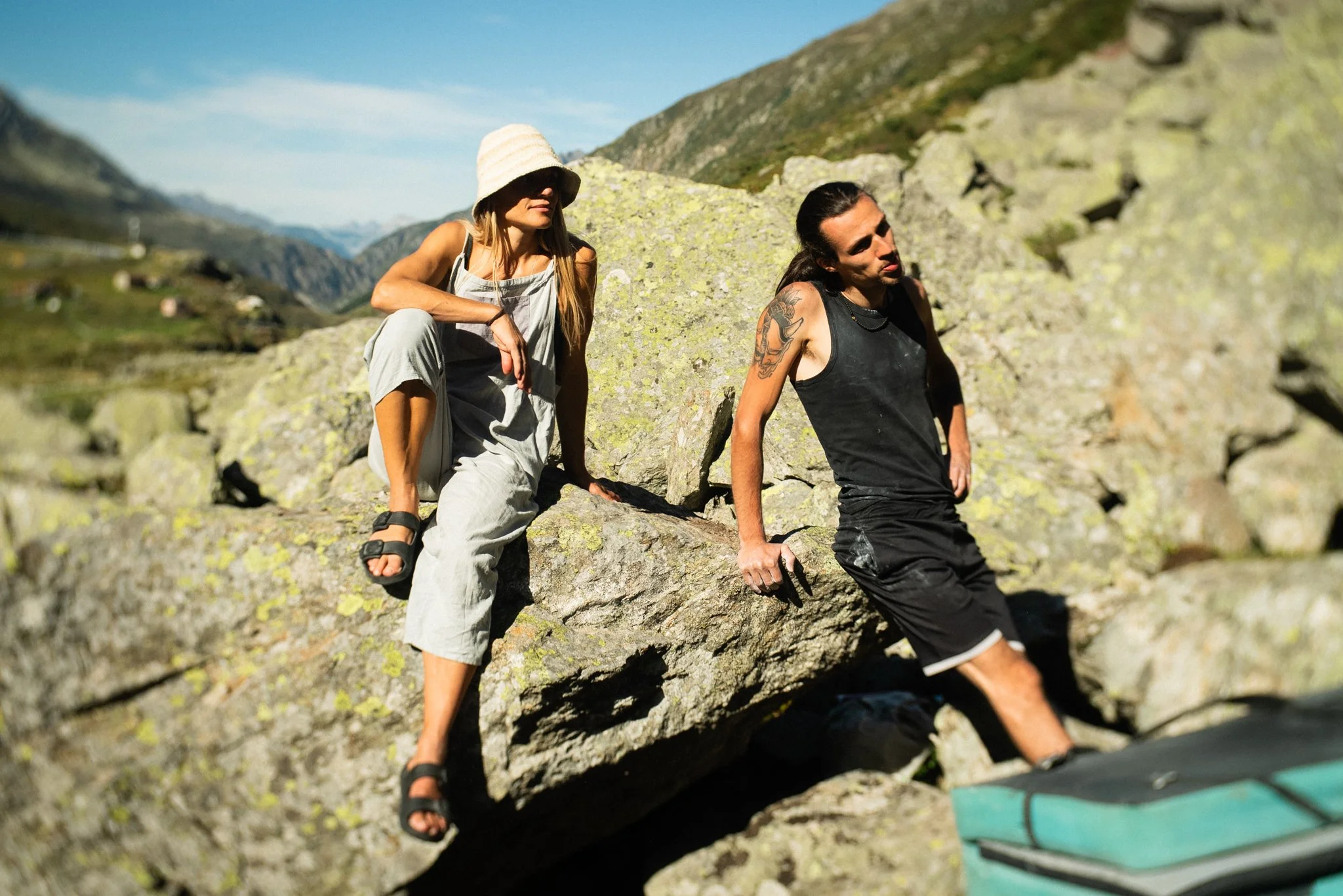 Two people with long dark hair, one woman wearing a sun hat and overalls, and one man with tattoos wearing a black sleeveless shirt, sitting on rocks in a mountainous outdoor setting.