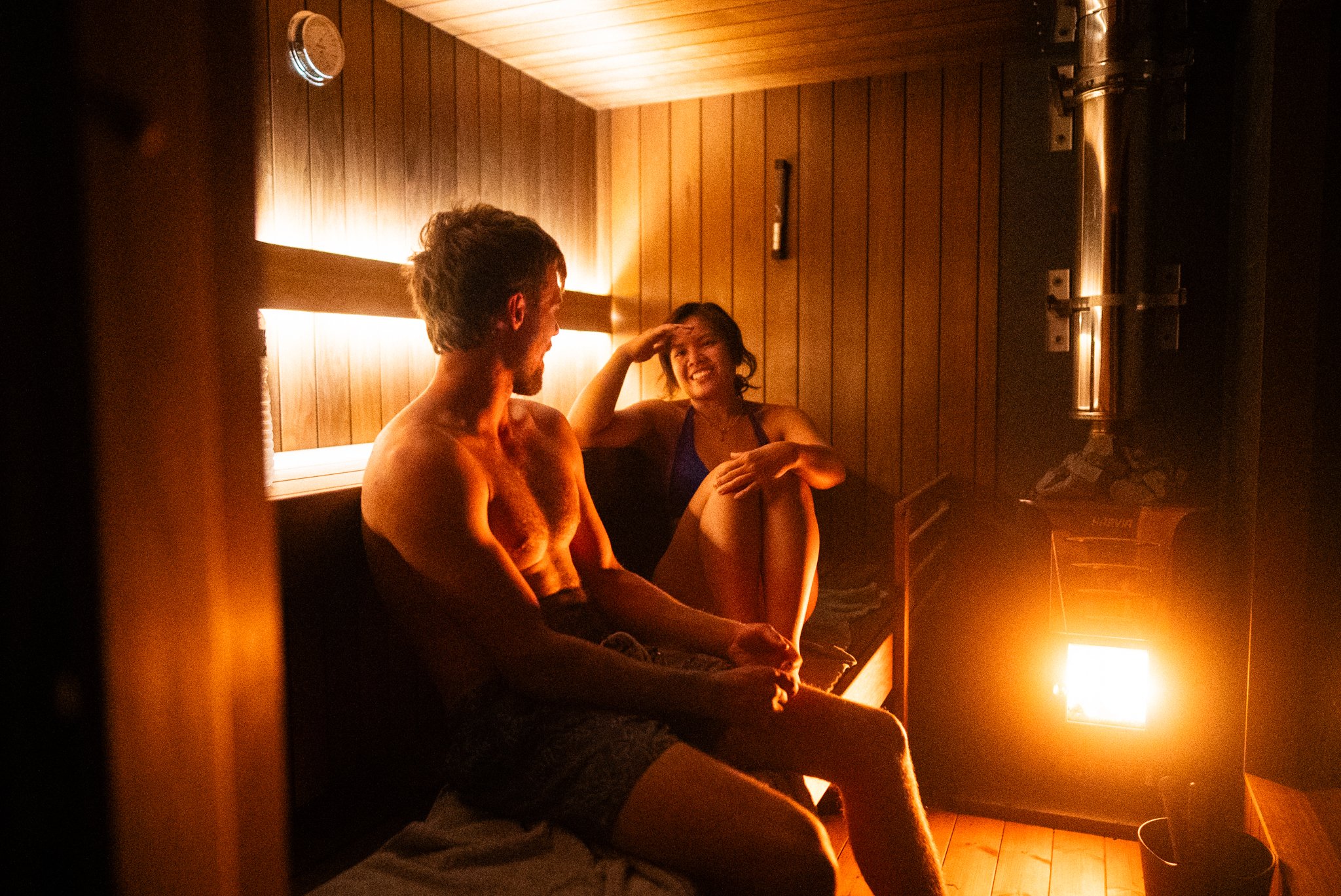 A man and woman sitting in a cozy wooden sauna, illuminated by a warm heater, smiling and engaging in conversation.
