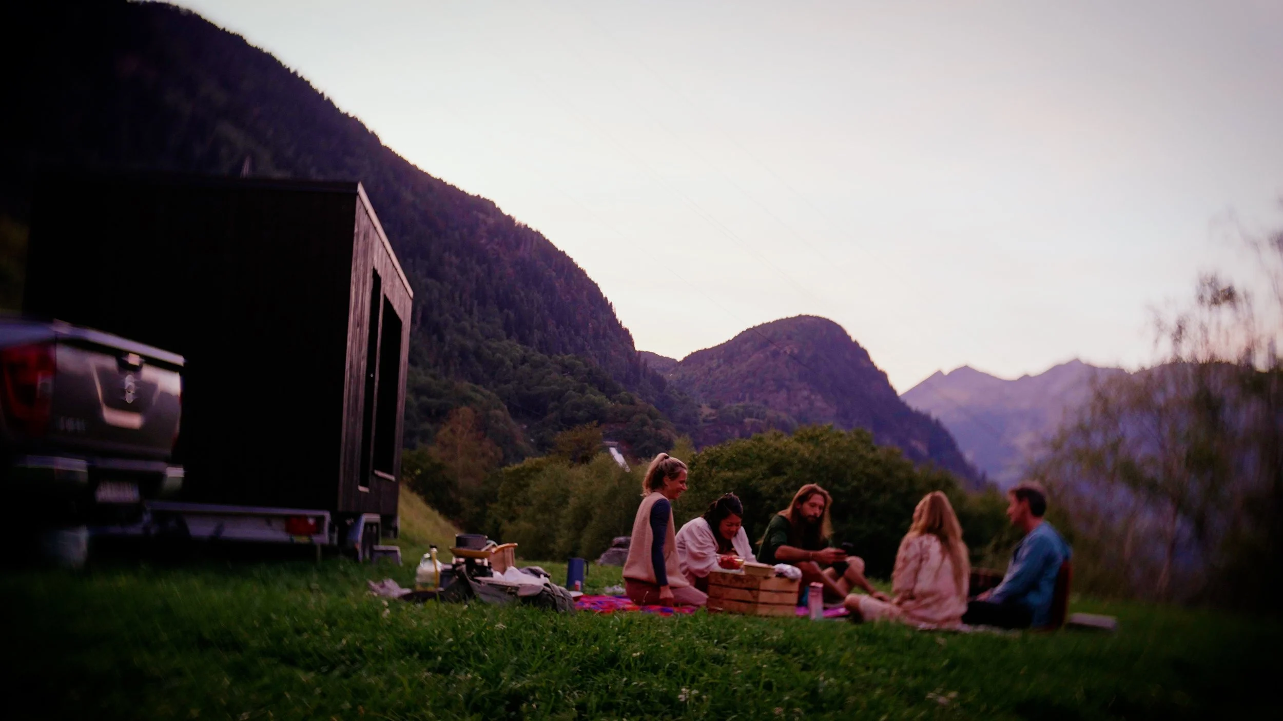People gathered outdoors on a grassy area during sunset, with mountains in the background, sitting around picnic items and socializing. In the background a mobile sauna.