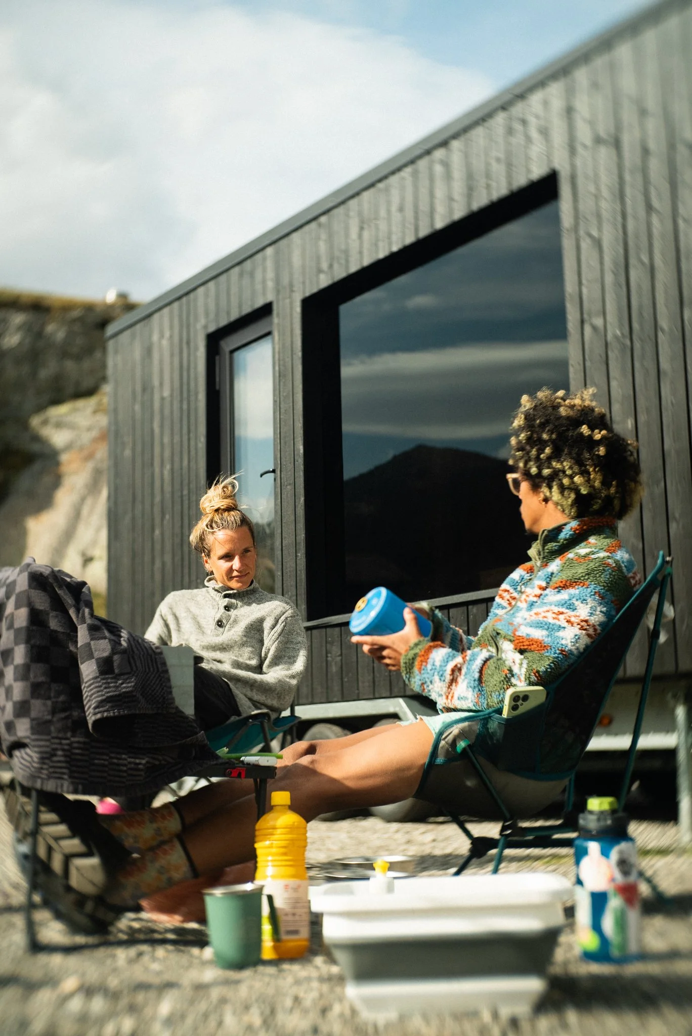 Two people sitting outside, one with curly hair and glasses, wearing a colorful fleece jacket, holding a blue container, and the other with her hair in a bun, wearing a gray sweater, sitting in front of a mobile sauna with large windows.