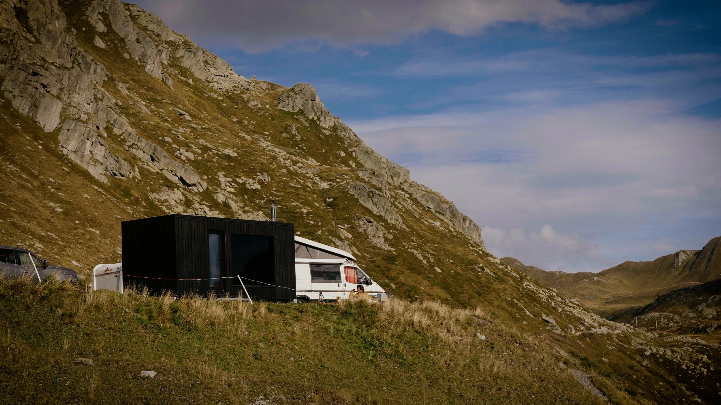 A white camper van and sauna house on grassy terrain, mountains in the background, and a cloudy sky.
