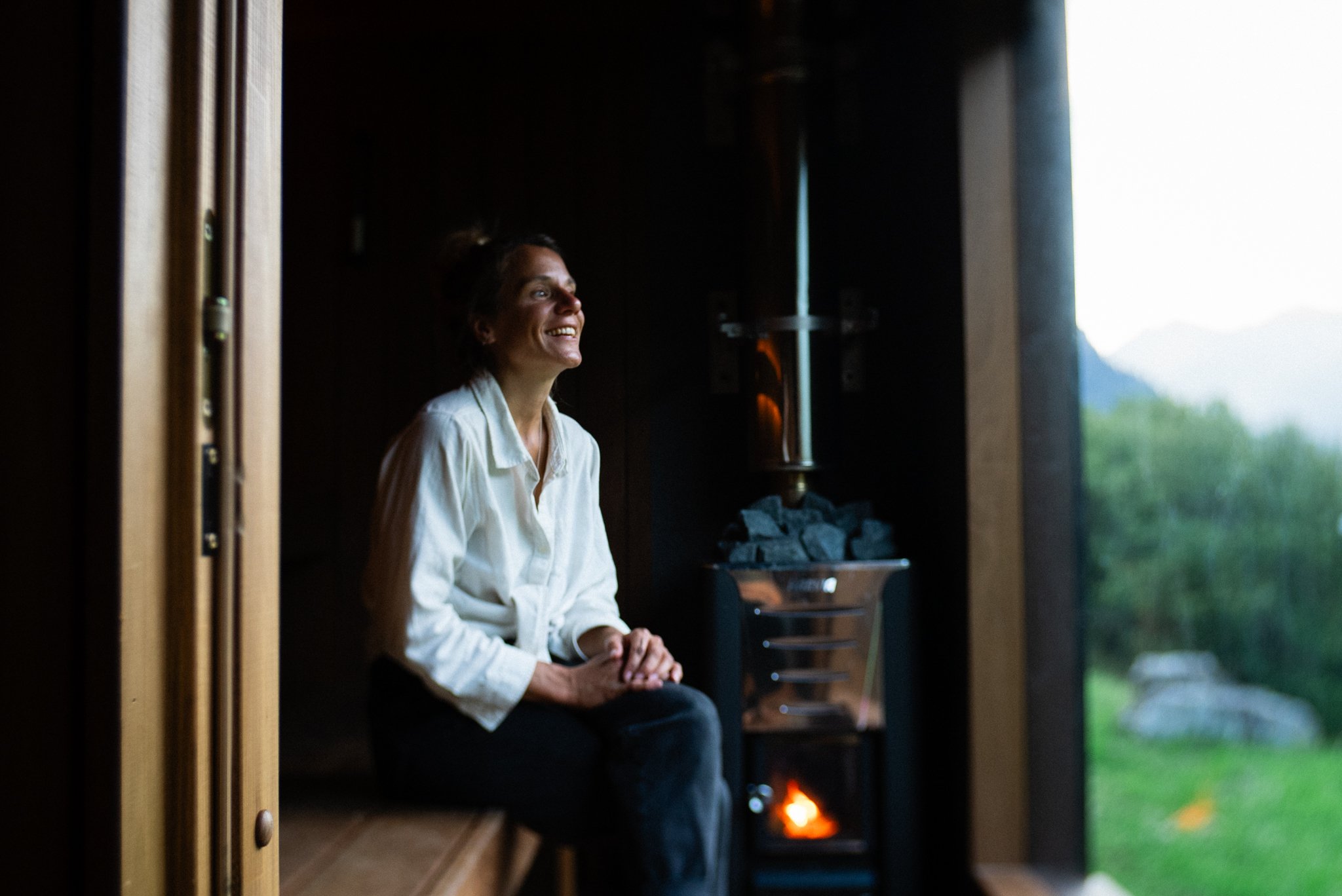 A woman sitting in a mobile sauna near the panoramic window, smiling and looking outside.