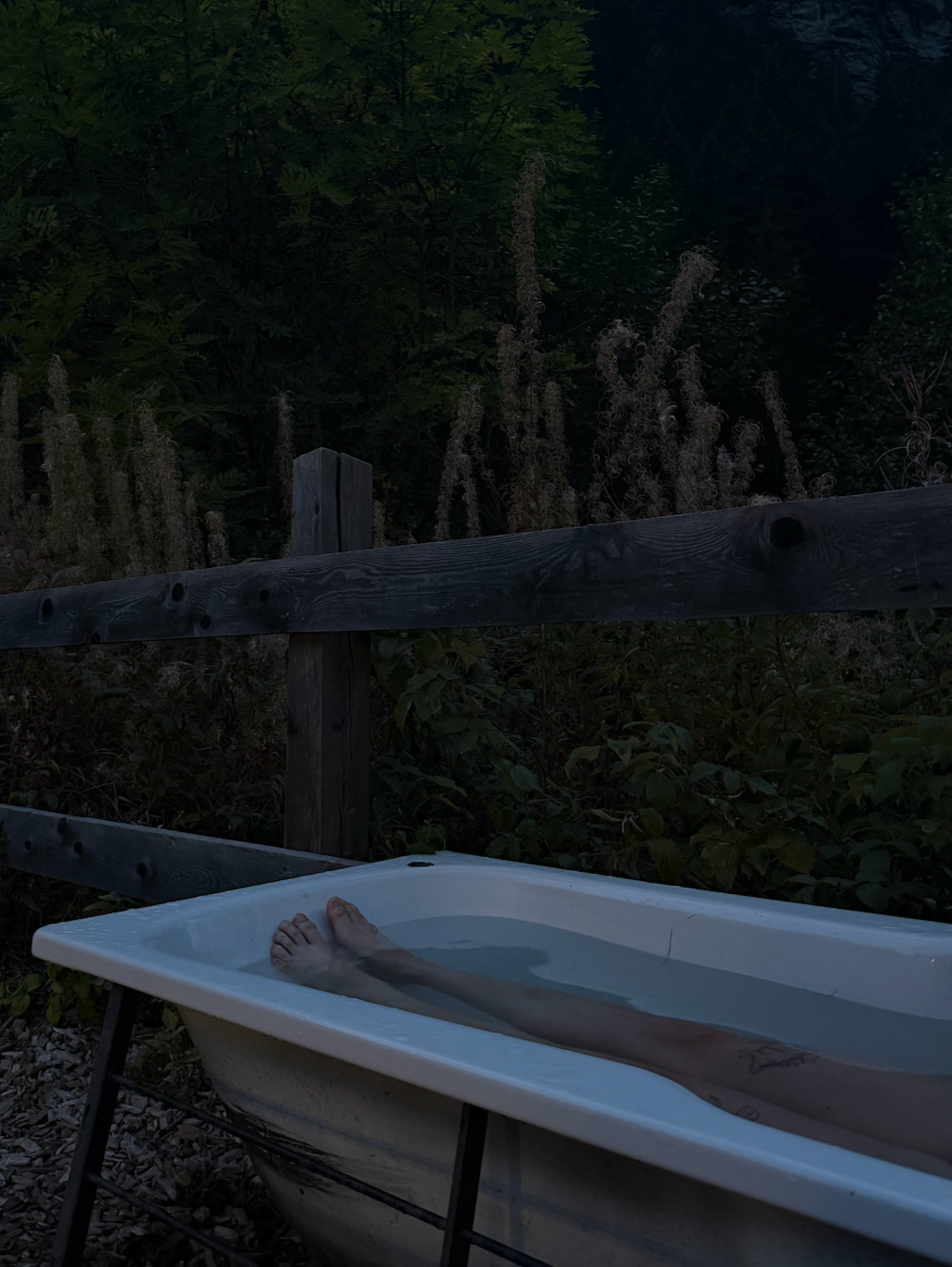 Feet submerged in an outdoor bathtub filled with water, with a wooden fence and dark green foliage in the background.