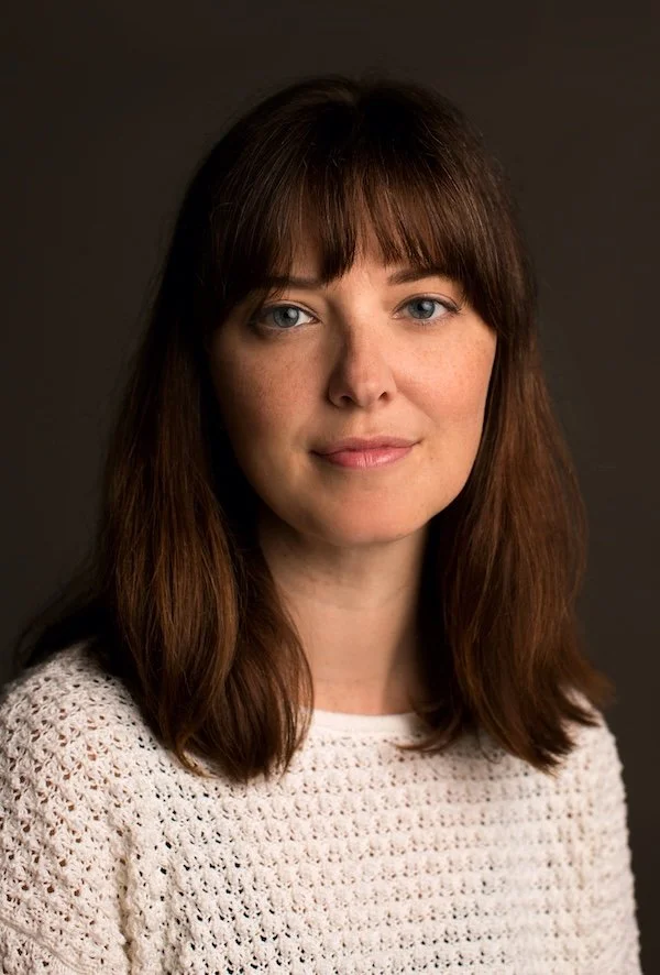 The author Jenn Thorne - a young woman with long brown hair and blue eyes, wearing a white knitted sweater against a dark background.