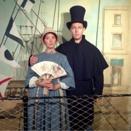 Two people dressed in historical costumes standing on a ship set with a painted background of buildings and flag.