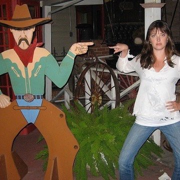 A woman posing next to a life-sized cartoon cutout of a cowboy, both pointing finger guns at each other, outside a building with a wagon wheel.