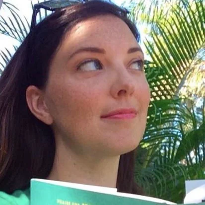 A woman with dark hair looking to the side, holding a green book outdoors with palm leaves in the background.