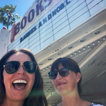 Two women smiling in front of a movie theater with a sign that reads 'BOOK' and parts of other words visible.