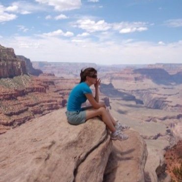 A young girl sitting on a rock ledge at the Grand Canyon, looking into the distance. She is wearing a blue shirt, shorts, and sneakers, with a cloudy sky overhead.