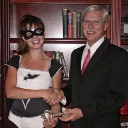 A woman wearing a black mask and a bat-themed dress shaking hands with a man in a suit, who is holding a key, in front of a bookshelf.