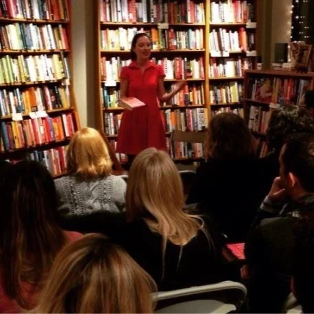 A woman in a red dress giving a talk or presentation in a bookstore to an audience of people seated in front of her.