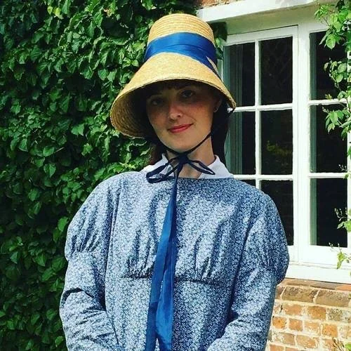 Woman wearing a wide-brimmed straw hat with a blue ribbon, blue patterned dress, standing in front of a brick wall and green leafy background.